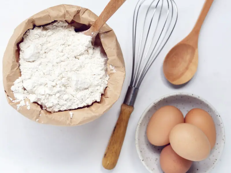 Batidor, huevos, cuchara de madera y harina para pasteles en una bolsa de papel 